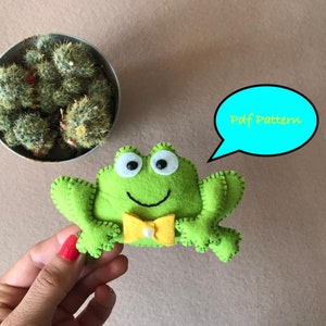 Könnte beinhalten: Grüner Filz-Frosch mit einer gelben Schleife und einem weißen Perlenknopf. Der Frosch hält eine Sprechblase, auf der "Pdf Pattern" steht.