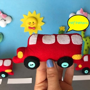 Op de afbeelding: Een rode bus van vilt met witte ramen en gele details. De bus heeft zwarte wielen met blauwe knoppen. De bus wordt vastgehouden door een hand. De achtergrond bevat een blauwe lucht, een gele zon, een witte wolk en twee groene cactussen. De tekst "Pdf Pattern" staat in een gele spreekballon.