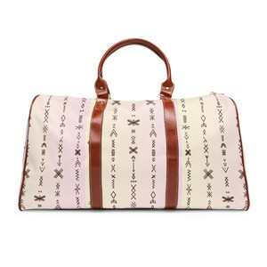 Könnte beinhalten: Eine cremefarbene Reisetasche mit einem geometrischen Muster in Braun. Die Tasche hat braune Ledergriffe und Akzente. Das Design umfasst vertikale Streifen und verschiedene Pfeil- und Kreuzformen. Die Tasche ist für Reisen konzipiert.