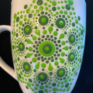 May include: A white ceramic mug with a green mandala design painted on the front. The design features a series of concentric circles and dots in shades of green.