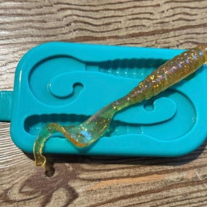May include: A turquoise silicone mold with two cavities shaped like a fish tail. A translucent yellow and gold glitter lure is in the mold.