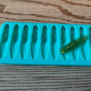 May include: A turquoise silicone mold with 12 cavities shaped like small fish. A green and gold glitter lure is sitting on top of the mold.