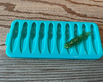 May include: A turquoise silicone mold with 12 cavities shaped like small fish. A green and gold glitter lure is sitting on top of the mold.