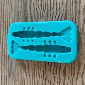 Puede incluir: Un molde de silicona turquesa con dos cavidades con forma de camarones. El molde está diseñado para hacer golosinas comestibles con forma de camarones.