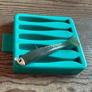 Könnte beinhalten: Ein dunkelgrüner Angelköder mit einem silbernen Haken liegt auf einem türkisfarbenen Kunststoff-Köderhalter. Der Köder hat einen hellgrünen Schwanz. Der Halter hat mehrere geformte Schlitze zur Aufbewahrung von Ködern. Der Hintergrund ist eine Holzoberfläche.