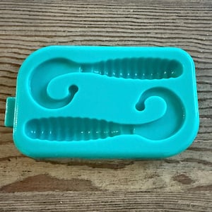 Puede incluir: Un molde de silicona azul turquesa para hacer dos señuelos de pesca con forma de gusano. El molde tiene dos cavidades, cada una con forma de gusano con una cola rizada.