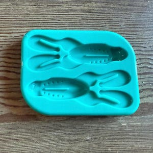 May include: A teal silicone mold with two cavities, each shaped like a stylized frog. The frog shapes have detailed features, including eyes and leg outlines. The mold is set against a wooden background.