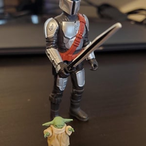 May include: A Mandalorian action figure with a silver helmet, armor, and a red strap, holding a black sword. A small green Grogu figure stands nearby, wearing a tan robe. The figures are on a dark surface.
