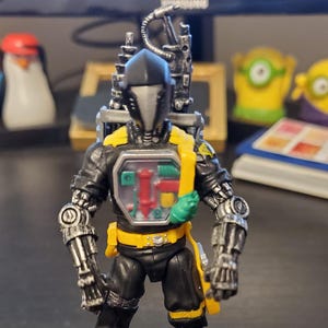 May include: A black and yellow action figure of a Cobra B.A.T. robot, with a clear chest revealing internal components. The figure has a black helmet, yellow boots, and a backpack. The base reads "CODE NAME: COBRA B.A.T."