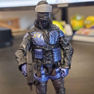 May include: A Snake Eyes action figure from the G.I. Joe franchise. The figure is dressed in a black and blue tactical suit, with a masked face and a visor. The figure is holding a weapon and stands on a base that reads "G.I. Joe" and "Code Name: Snake Eyes."