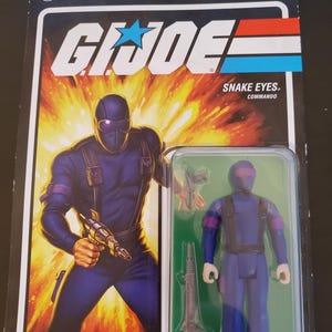 May include: A G.I. Joe Snake Eyes Commando action figure in its original packaging. The figure is in a clear plastic case, with a purple and black color scheme. The packaging features the G.I. Joe logo and the text "Snake Eyes. Commando."
