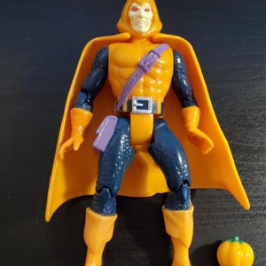May include: A yellow and blue action figure of a superhero wearing a yellow cape and a purple belt. The figure has a pumpkin accessory.