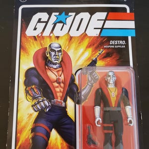 May include: A G.I. Joe Destro action figure in its original packaging. The card features a dynamic illustration of Destro with a weapon, and the figure is visible in a clear plastic bubble. The packaging includes the text "ReAction Figures" and "Destro, Weapons Supplier."