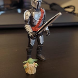 May include: A Mandalorian action figure in silver and black armor, holding a black sword with a silver handle. A small green figure of Grogu, wearing a tan robe, stands nearby. The figures are on a dark surface.