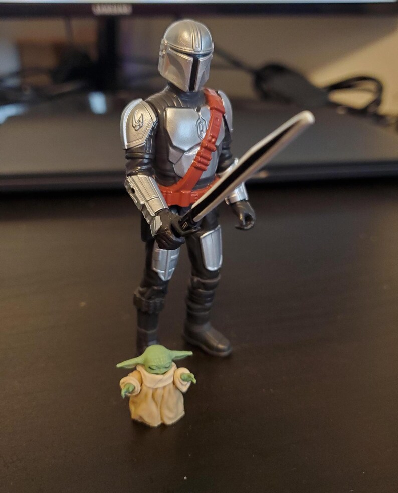 May include: A Mandalorian action figure in silver and black armor, holding a black sword with a silver handle. A small green figure of Grogu, wearing a tan robe, stands nearby. The figures are on a dark surface.
