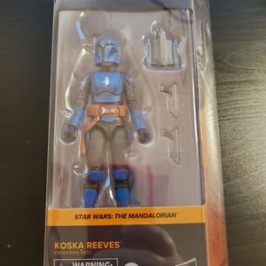 May include: A blue and gray action figure of Koska Reeves from Star Wars: The Mandalorian. The figure is packaged in a clear plastic box with a black and orange background. The box has the Star Wars logo and the words "The Black Series" on it. The figure is holding a blaster rifle in its right hand. The figure is wearing a blue helmet and armor. The figure is standing on a black base. The box also includes a warning label that says "Choking Hazard - Small parts. Not for children under 3 years."