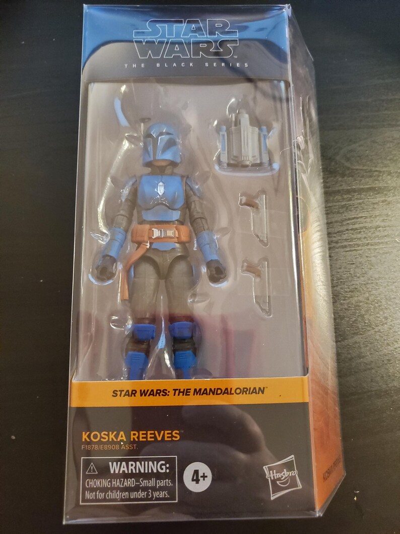May include: A blue and gray action figure of Koska Reeves from Star Wars: The Mandalorian. The figure is packaged in a clear plastic box with a black and orange background. The box has the Star Wars logo and the words "The Black Series" on it. The figure is holding a blaster rifle in its right hand. The figure is wearing a blue helmet and armor. The figure is standing on a black base. The box also includes a warning label that says "Choking Hazard - Small parts. Not for children under 3 years."