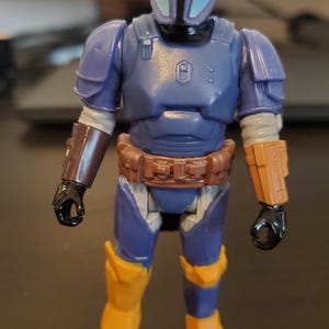 May include: A blue and yellow action figure of a character in armor. The figure has a helmet with a T-shaped visor, a brown belt, and yellow accents on the legs and arms. The figure is standing upright.