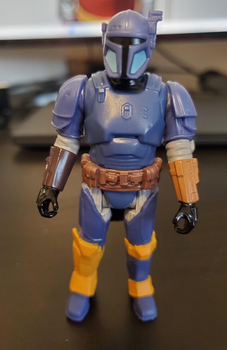 May include: A blue and yellow action figure of a character in armor. The figure has a helmet with a T-shaped visor, a brown belt, and yellow accents on the legs and arms. The figure is standing upright.