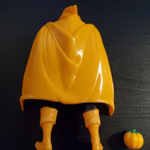 May include: A yellow plastic action figure with a cape and orange boots. The figure is standing on a dark brown surface. A small orange pumpkin is on the right side of the image.