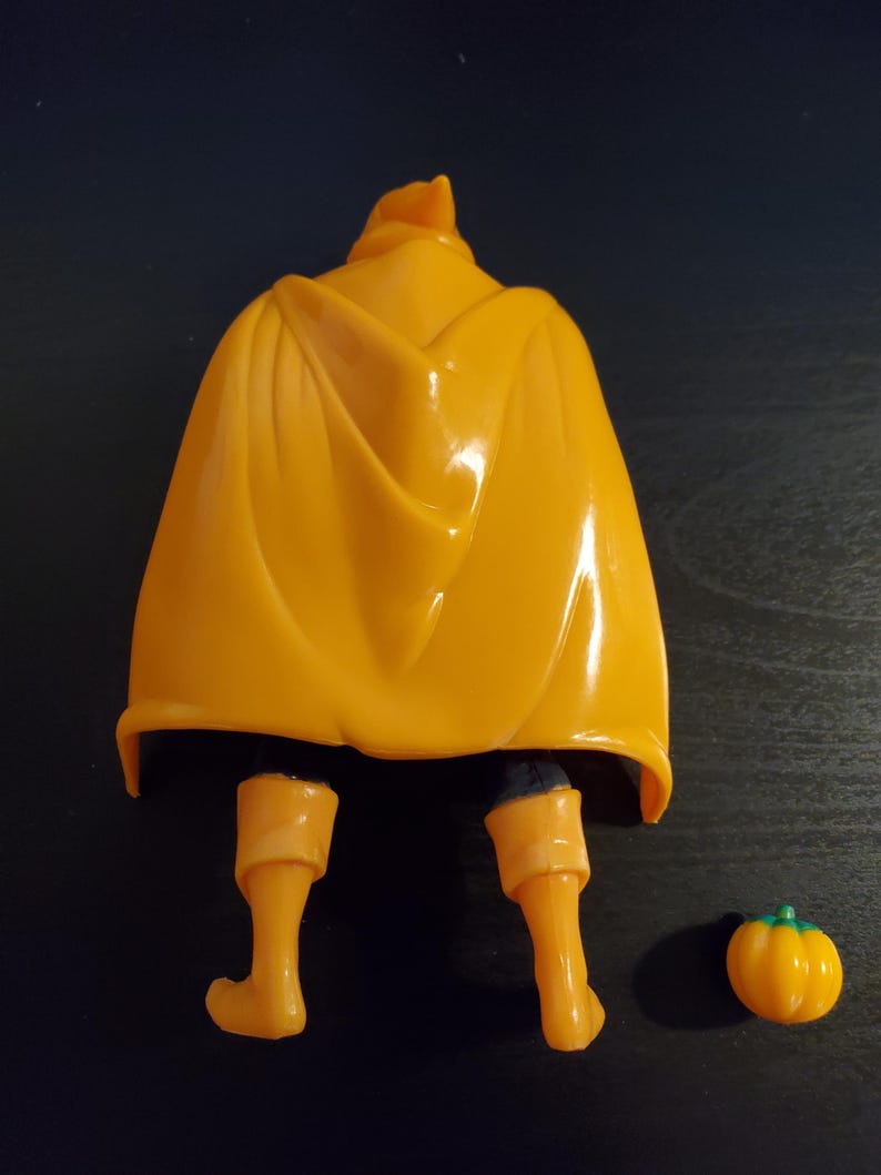 May include: A yellow plastic action figure with a cape and orange boots. The figure is standing on a dark brown surface. A small orange pumpkin is on the right side of the image.