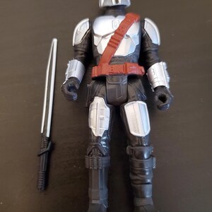 May include: A Mandalorian action figure with a silver helmet, chest armor, and black suit. The figure has a red belt and shoulder strap. A silver and black sword is placed next to the figure. The toy is a collectible item.