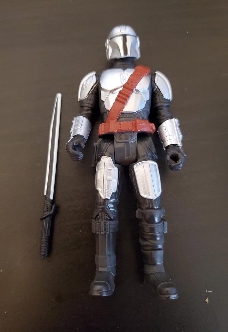 May include: A Mandalorian action figure with a silver helmet, chest armor, and black suit. The figure has a red belt and shoulder strap. A silver and black sword is placed next to the figure. The toy is a collectible item.