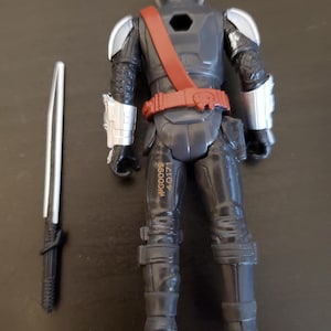 May include: A black action figure with silver accents, a red belt, and a silver helmet. The figure is holding a silver and black sword. The figure has the text #G0099 4-0171 printed on its leg.