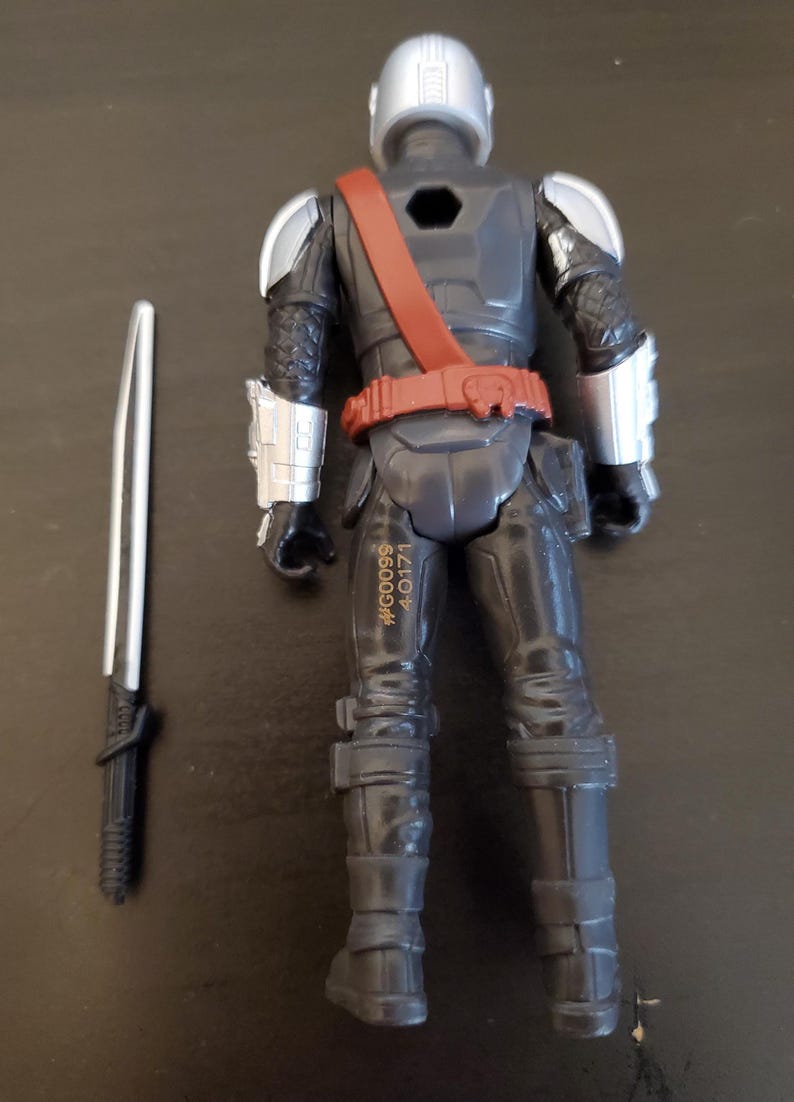 May include: A black action figure with silver accents, a red belt, and a silver helmet. The figure is holding a silver and black sword. The figure has the text #G0099 4-0171 printed on its leg.