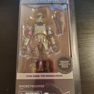 May include: A Star Wars: The Mandalorian Shore Trooper action figure in a plastic package. The figure is wearing gold armor with a red stripe on the left shoulder. The figure is holding a blaster rifle. The package has a warning label for children under 3 years old.