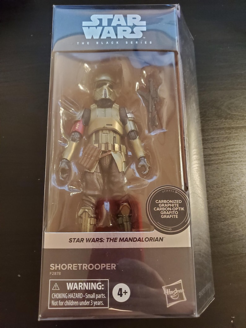 May include: A Star Wars: The Mandalorian Shore Trooper action figure in a plastic package. The figure is wearing gold armor with a red stripe on the left shoulder. The figure is holding a blaster rifle. The package has a warning label for children under 3 years old.