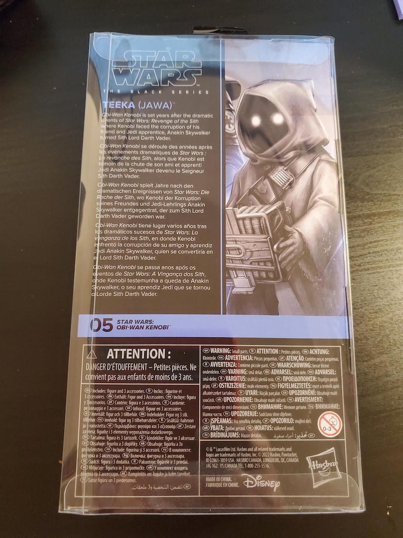 May include: A Star Wars: The Black Series action figure of Teeka, a Jawa from the Star Wars universe. The figure is packaged in a clear plastic box with a black and white background. The box features a photo of Teeka and text about the character. The text on the box reads "Star Wars: The Black Series Teeka (Jawa)".