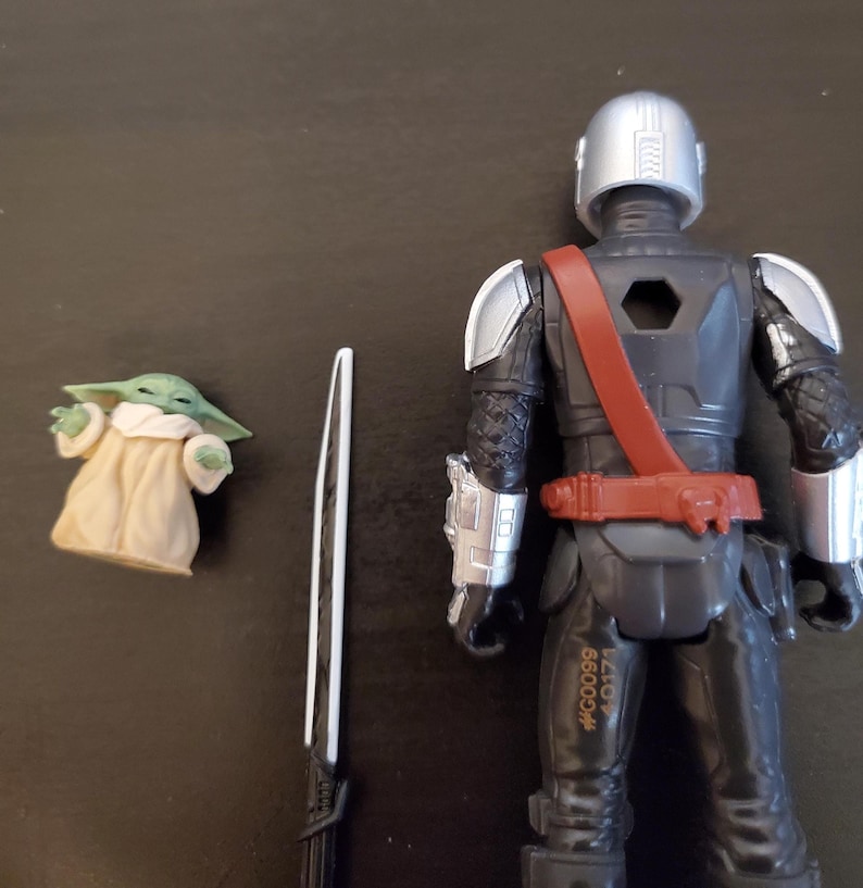 May include: Action figure set featuring a Mandalorian-style character in black and silver armor, a small green alien figure in a tan robe, and a black and silver sword. The Mandalorian figure has a red belt and shoulder strap. The set is a collectible toy.