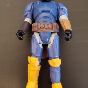 May include: A blue and yellow action figure of a character in armor. The figure has a helmet, chest armor, and leg armor. The figure has a brown belt and black gloves. The figure is standing on a dark surface.