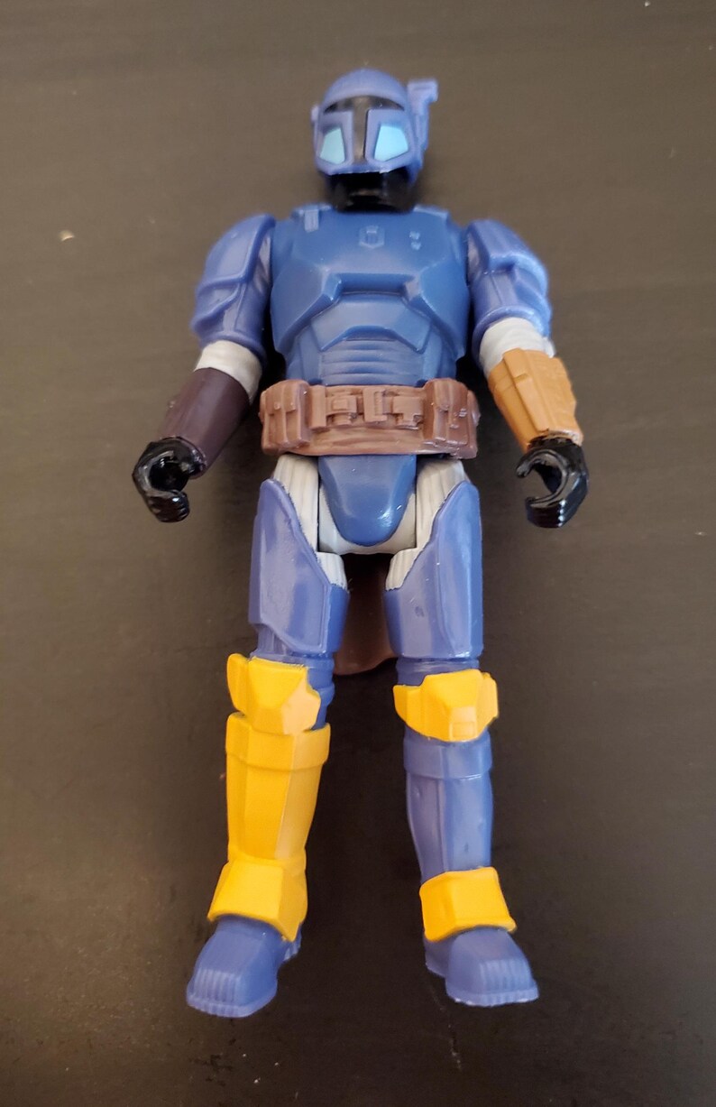 May include: A blue and yellow action figure of a character in armor. The figure has a helmet, chest armor, and leg armor. The figure has a brown belt and black gloves. The figure is standing on a dark surface.