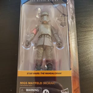 May include: A Star Wars: The Mandalorian action figure of Migs Mayfield (Morak) in a gray and black uniform with a red armband. The figure is packaged in a clear plastic box with a black and orange background. The box has the Star Wars logo and the words "The Black Series" on it. The figure is recommended for ages 4 and up.