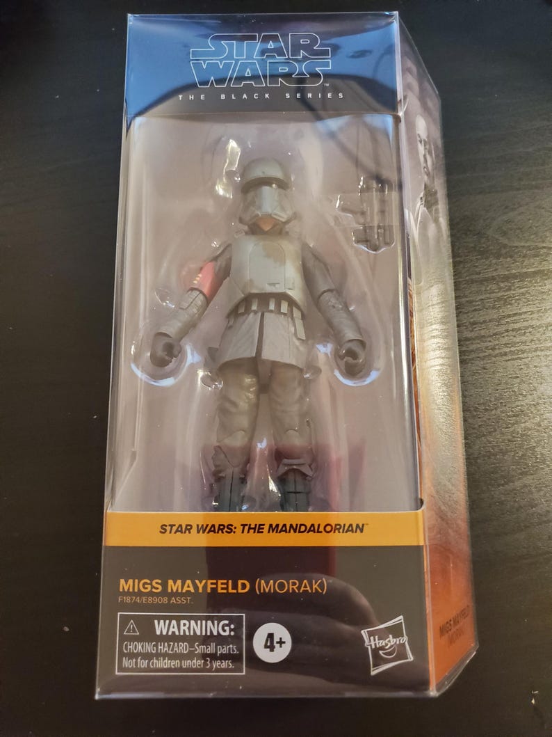 May include: A Star Wars: The Mandalorian action figure of Migs Mayfield (Morak) in a gray and black uniform with a red armband. The figure is packaged in a clear plastic box with a black and orange background. The box has the Star Wars logo and the words "The Black Series" on it. The figure is recommended for ages 4 and up.