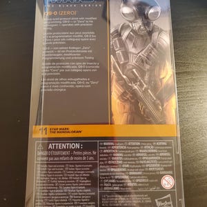 May include: A black and silver Star Wars: The Mandalorian action figure of a protocol droid with large bug eyes. The figure is packaged in a clear plastic box with a black and yellow background. The text on the box reads "Star Wars: The Black Series" and "09-0 (Zero)".