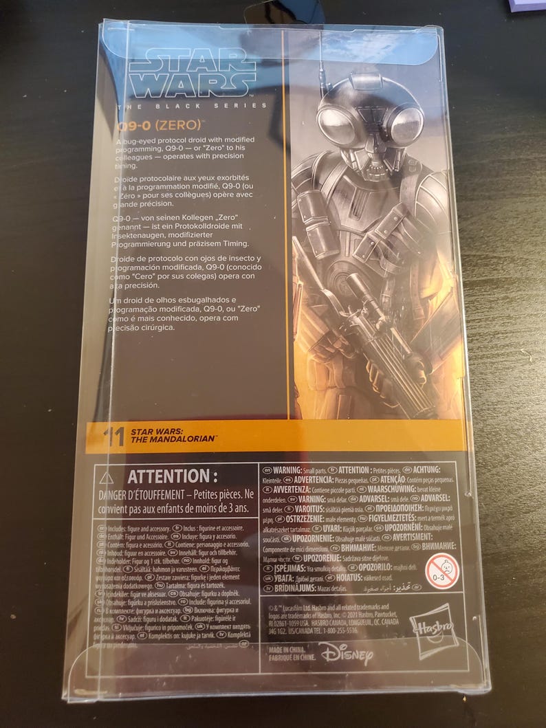 May include: A black and silver Star Wars: The Mandalorian action figure of a protocol droid with large bug eyes. The figure is packaged in a clear plastic box with a black and yellow background. The text on the box reads "Star Wars: The Black Series" and "09-0 (Zero)".