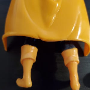 May include: Close-up of the lower legs of an action figure. The figure is wearing yellow pants and boots. The figure is wearing a yellow cape that is draped over the figure's legs.