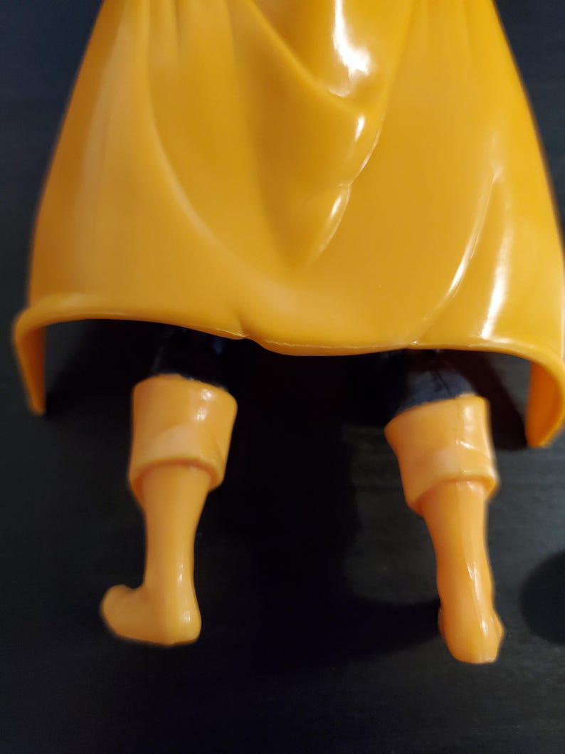 May include: Close-up of the lower legs of an action figure. The figure is wearing yellow pants and boots. The figure is wearing a yellow cape that is draped over the figure's legs.