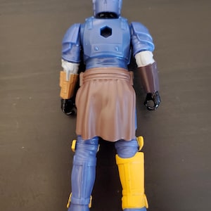 May include: A blue action figure with a brown skirt and yellow boots. The figure has a helmet and armor, with brown and gold accents. The figure is standing on a dark surface.