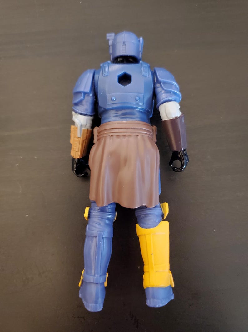 May include: A blue action figure with a brown skirt and yellow boots. The figure has a helmet and armor, with brown and gold accents. The figure is standing on a dark surface.