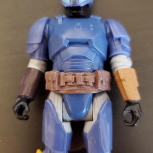 May include: A blue action figure with a helmet and armor, featuring brown and yellow accents. The figure has a brown belt and arm guards, with black hands and feet. The figure is approximately 6 inches tall.