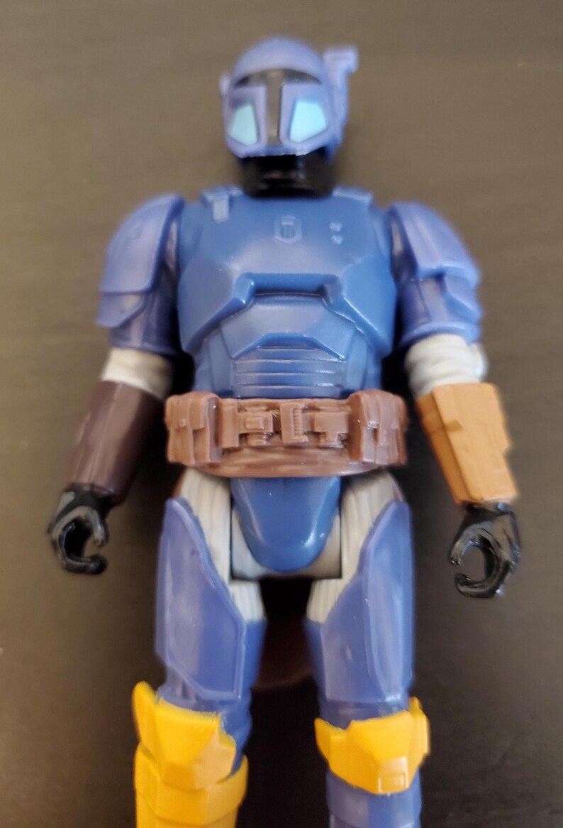 May include: A blue action figure with a helmet and armor, featuring brown and yellow accents. The figure has a brown belt and arm guards, with black hands and feet. The figure is approximately 6 inches tall.