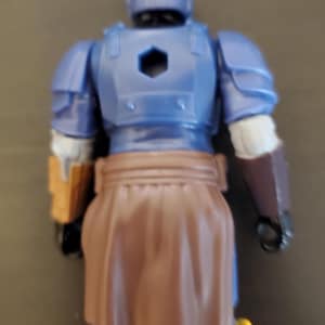 May include: A blue action figure of a warrior, viewed from the back. The figure has a helmet, armor, and a brown skirt-like garment. The figure has yellow and brown accents.