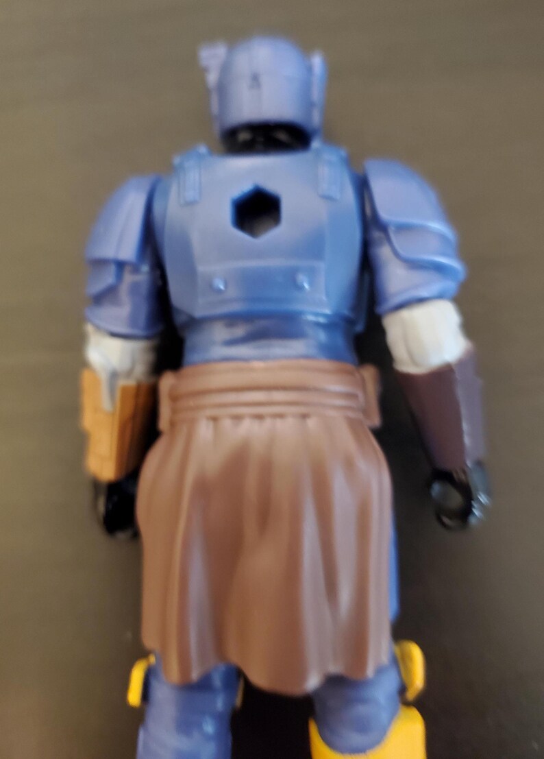 May include: A blue action figure of a warrior, viewed from the back. The figure has a helmet, armor, and a brown skirt-like garment. The figure has yellow and brown accents.