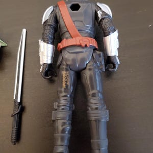 May include: A black action figure of a character in armor, with silver accents on the shoulders and helmet. The figure has a red belt and strap, and a silver and black sword is next to it. The figure has the text "WG0098 40171" printed on the leg.