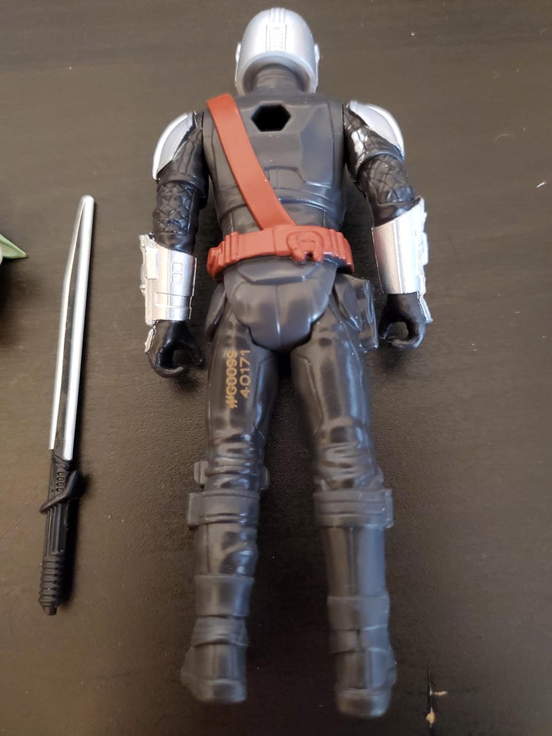 May include: A black action figure of a character in armor, with silver accents on the shoulders and helmet. The figure has a red belt and strap, and a silver and black sword is next to it. The figure has the text "WG0098 40171" printed on the leg.