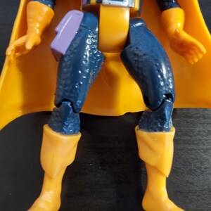 May include: A blue and yellow action figure with a purple sash and a yellow cape. The figure has a detailed design and is posed with its legs bent.